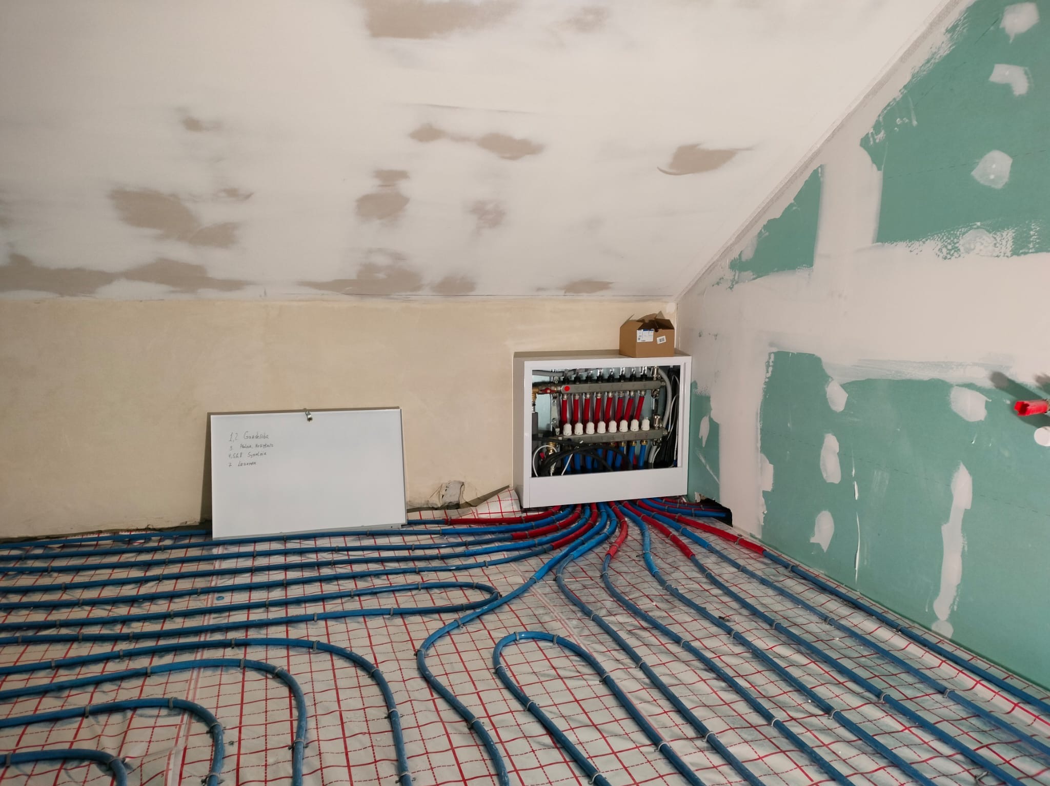 Instalacja ogrzewania podłogowego w poddaszu: rozłożone niebieskie rury na siatce, biała szafka rozdzielcza z czerwonymi elementami, ściany w trakcie wykończenia.