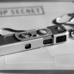 Czarno-białe ujęcie szpiegowskiej miniaturowej kamery z łańcuszkiem, leżącej na dokumentach oznaczonych jako 'TOP SECRET', obok spinacza do papieru.