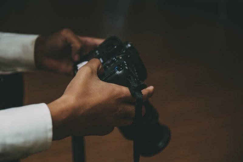 Dłonie mężczyzny w białej koszuli trzymają czarny aparat fotograficzny DSLR z paskiem, gotowy do zrobienia zdjęcia w słabym oświetleniu.