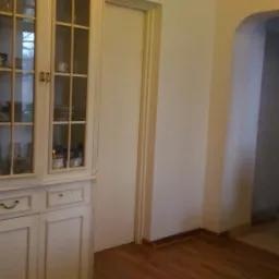 Fragment pokoju z białym, przeszklonym kredensem wypełnionym naczyniami, widoczne drzwi i łuk wejściowy do pomieszczenia wyłożonego szarymi płytkami.