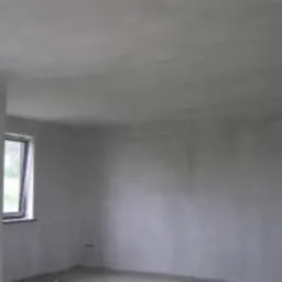 Szare, surowe wnętrze pokoju po nałożeniu świeżej warstwy gładzi na ściany i sufit, widoczne okno z zielenią za szybą.