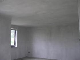 Szare, surowe wnętrze pokoju po nałożeniu świeżej warstwy gładzi na ściany i sufit, widoczne okno z zielenią za szybą.