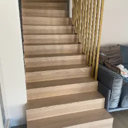 Jasne, drewniane schody z pionowymi, złotymi elementami balustrady w nowoczesnym wnętrzu, widok z dołu.