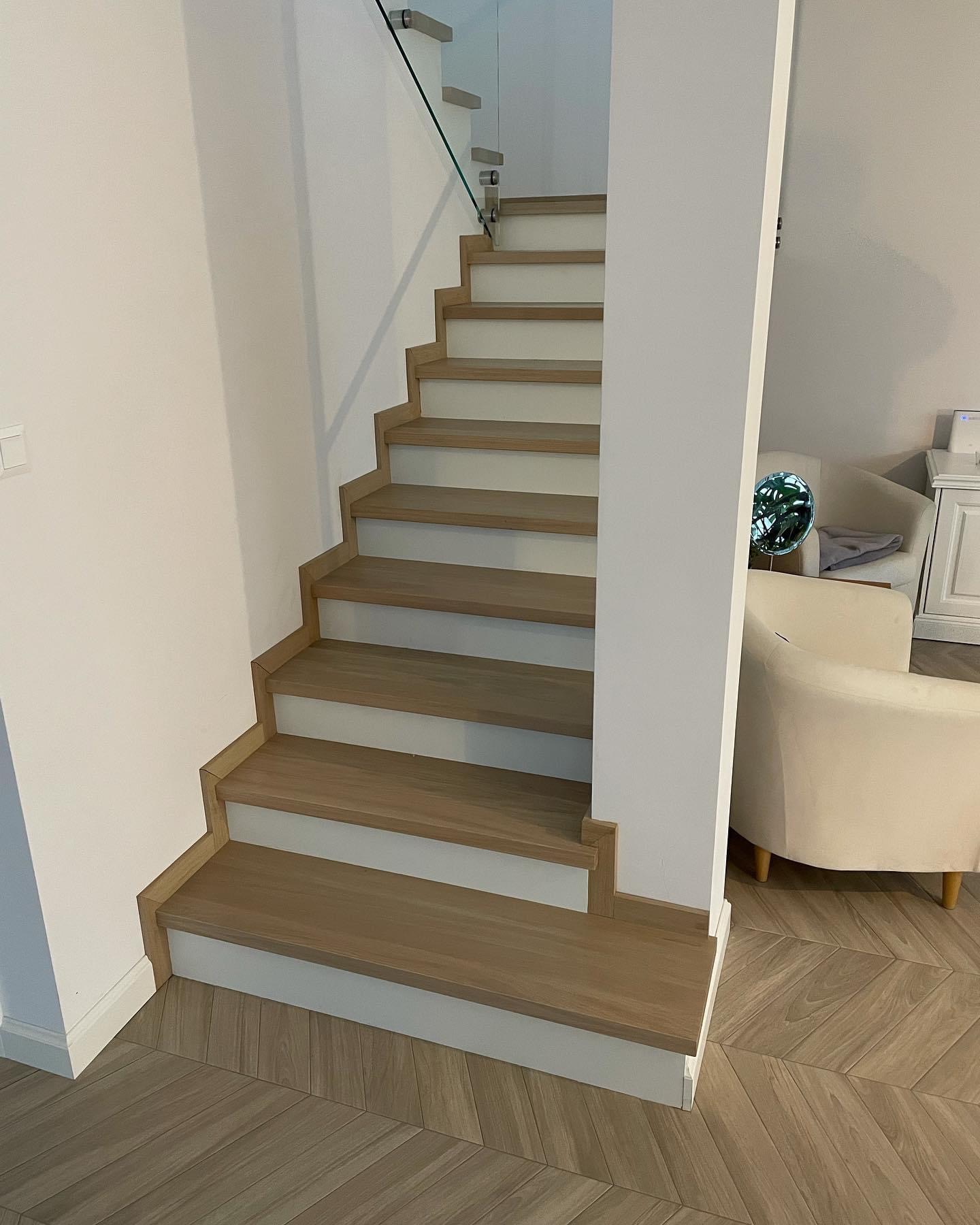 Drewniane schody z białymi podstopnicami i szklaną balustradą, widok z dołu, jasne wnętrze z parkietem w jodełkę.