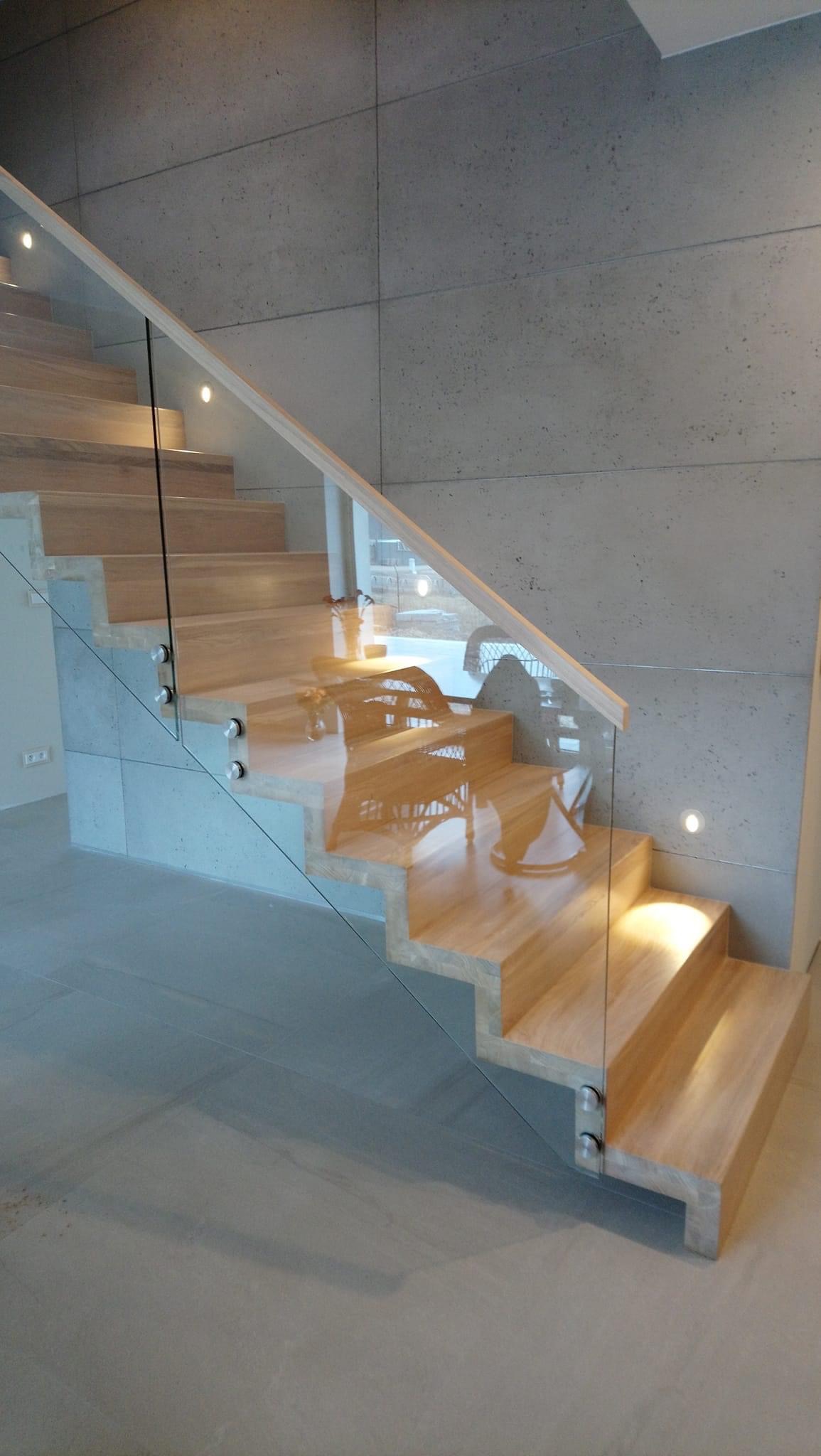 Nowoczesne schody z jasnego drewna z balustradą ze szkła mocowaną na metalowych uchwytach, oświetlenie punktowe w ścianie i stopniach, minimalistyczne wnętrze.