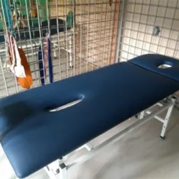 Ciemnoniebieski stół do masażu z otworem na twarz, ustawiony w pomieszczeniu z metalową siatką na ścianach i wiszącymi narzędziami.