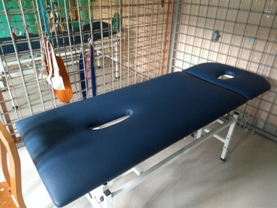 Ciemnoniebieski stół do masażu z otworem na twarz, ustawiony w pomieszczeniu z metalową siatką na ścianach i wiszącymi narzędziami.