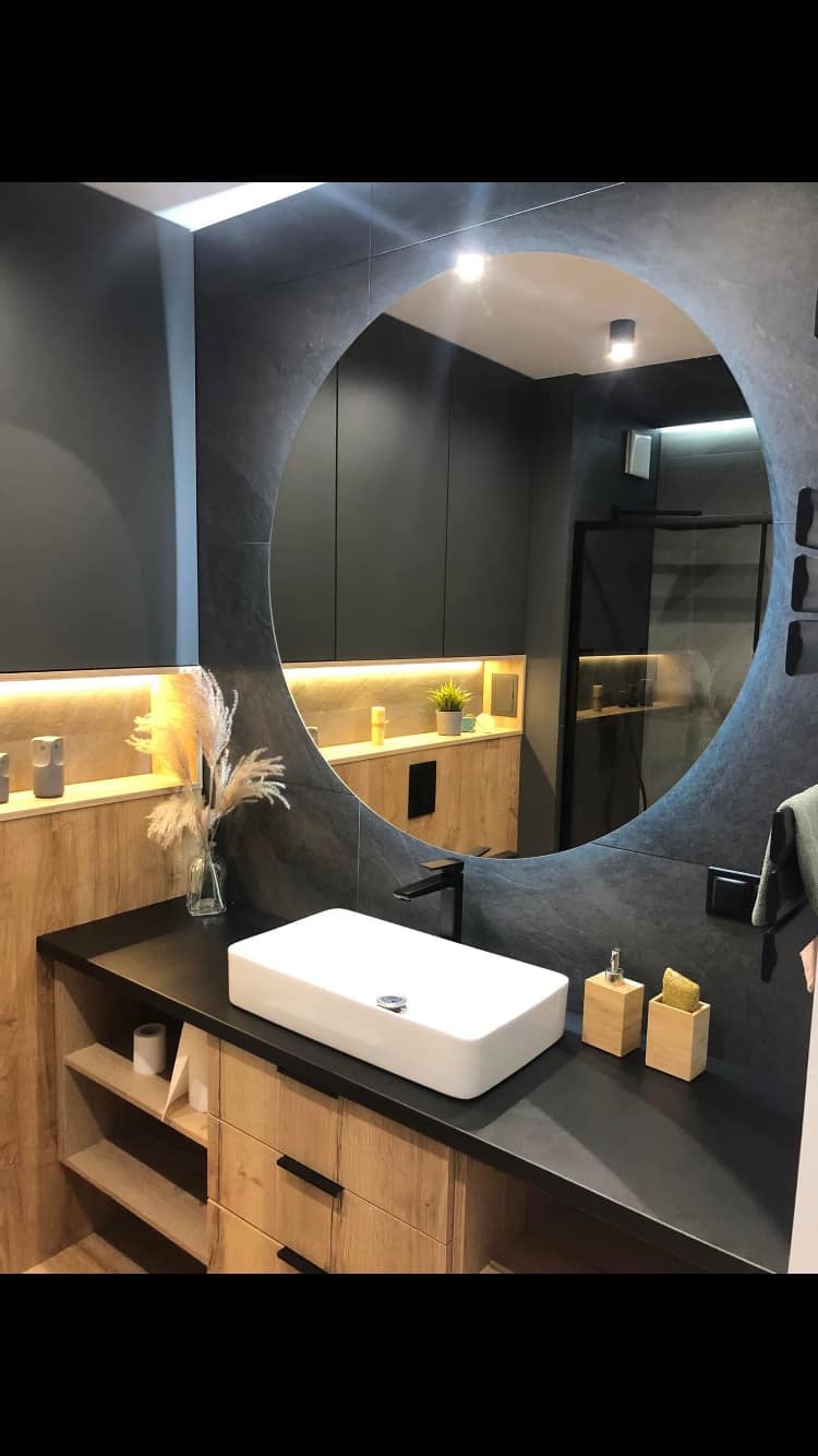 Nowoczesna łazienka z okrągłym lustrem, białą prostokątną umywalką nablatową, czarną armaturą i drewnianymi szafkami pod umywalką oraz dekoracyjną trawą pampasową w szklanym wazonie.