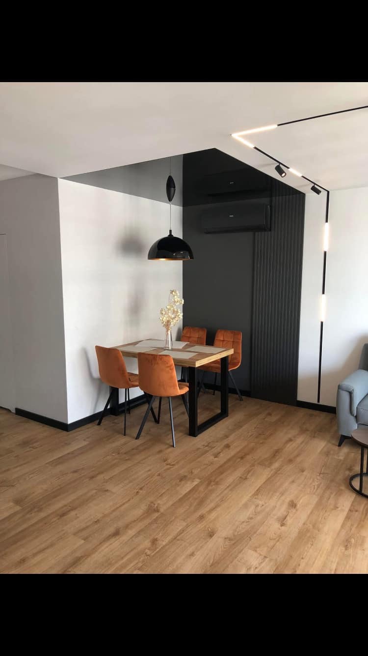Nowoczesna jadalnia z drewnianą podłogą, pomarańczowymi krzesłami i stołem z czarnymi nogami, oświetlona czarną lampą wiszącą i listwami LED na suficie.