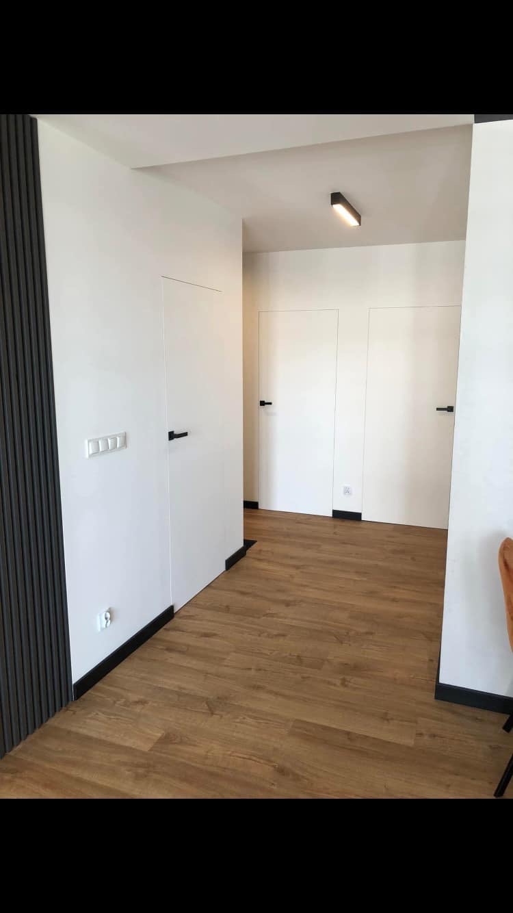 Nowoczesne wnętrze: drewnopodobna podłoga, białe ściany z ukrytymi drzwiami z czarnymi klamkami, dekoracyjny panel ścienny z pionowymi listwami.