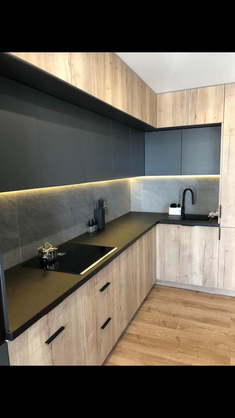 Nowoczesna kuchnia z szarymi szafkami górnymi i drewnopodobnymi dolnymi, czarnym blatem i armaturą, podświetleniem LED oraz płytkami imitującymi kamień na ścianie.