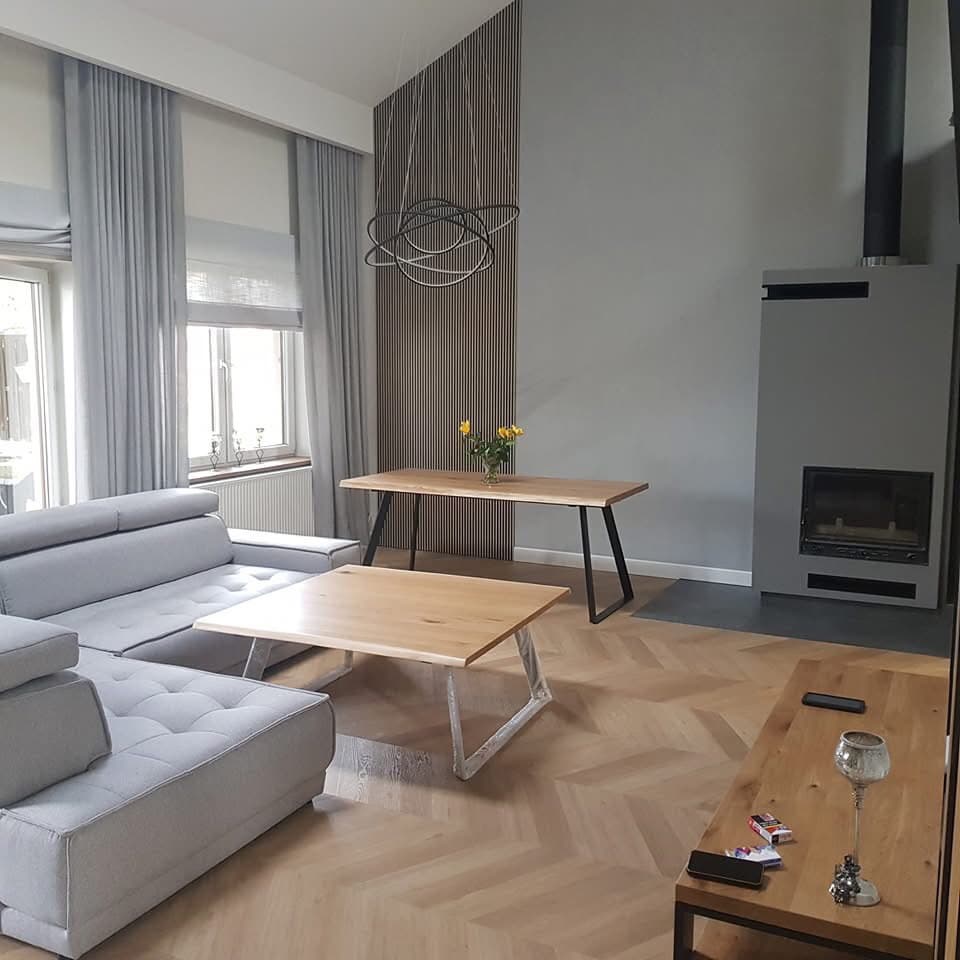 Jasny salon z parkietem ułożonym w jodełkę, szarą sofą, drewnianymi stolikami kawowymi na metalowych nogach, ścianą z drewnianych paneli i nowoczesnym kominkiem w szarej obudowie.