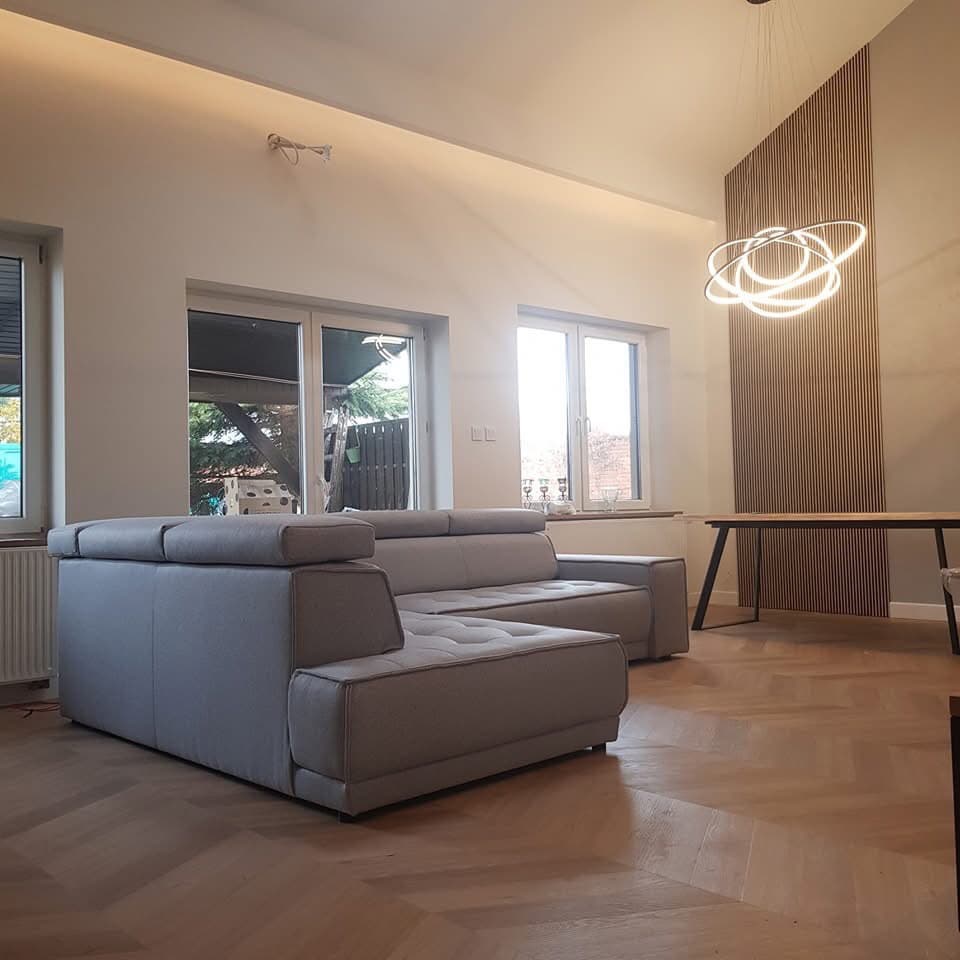 Jasny salon w Krzczonowie z szarą sofą narożną, parkietem ułożonym we wzór jodełki, drewnianą ścianą dekoracyjną i nowoczesną lampą wiszącą w kształcie pierścieni.