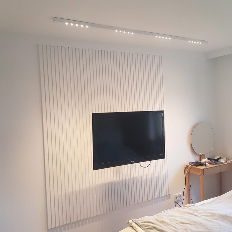 Biała ściana z pionowymi panelami, na której zawieszony jest telewizor, oświetlona liniowymi lampami LED na suficie, z fragmentem sypialni w tle.