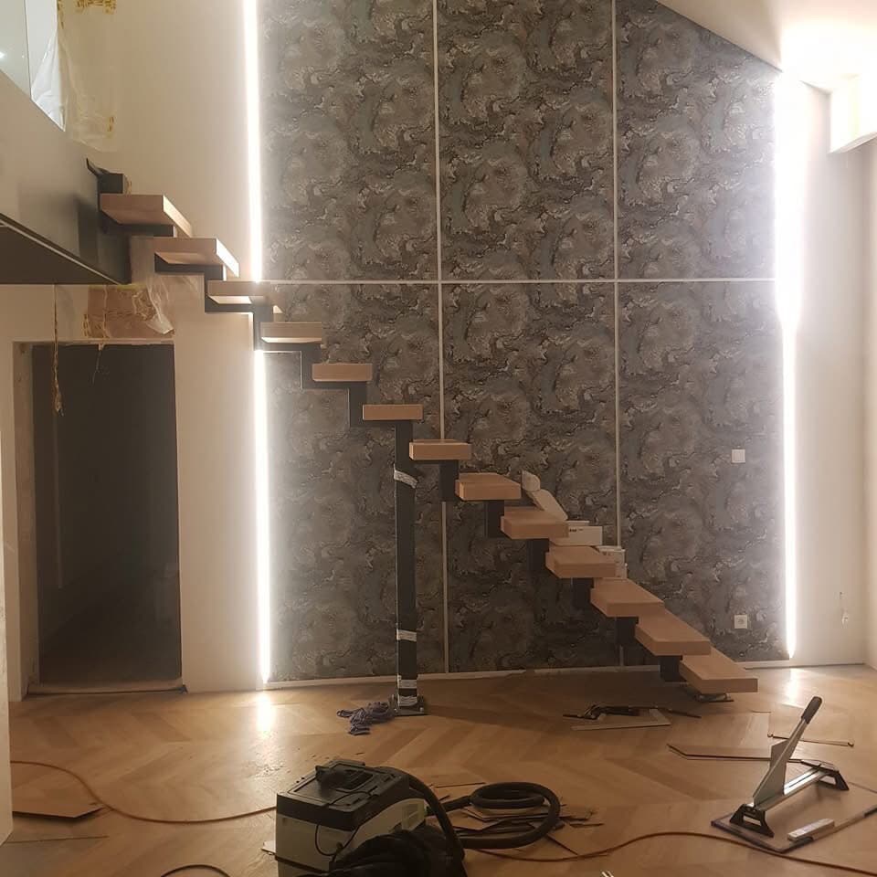 Montaż ażurowych schodów drewnianych na metalowej konstrukcji w pomieszczeniu z parkietem i wzorzystą tapetą, widoczne narzędzia i odkurzacz budowlany.