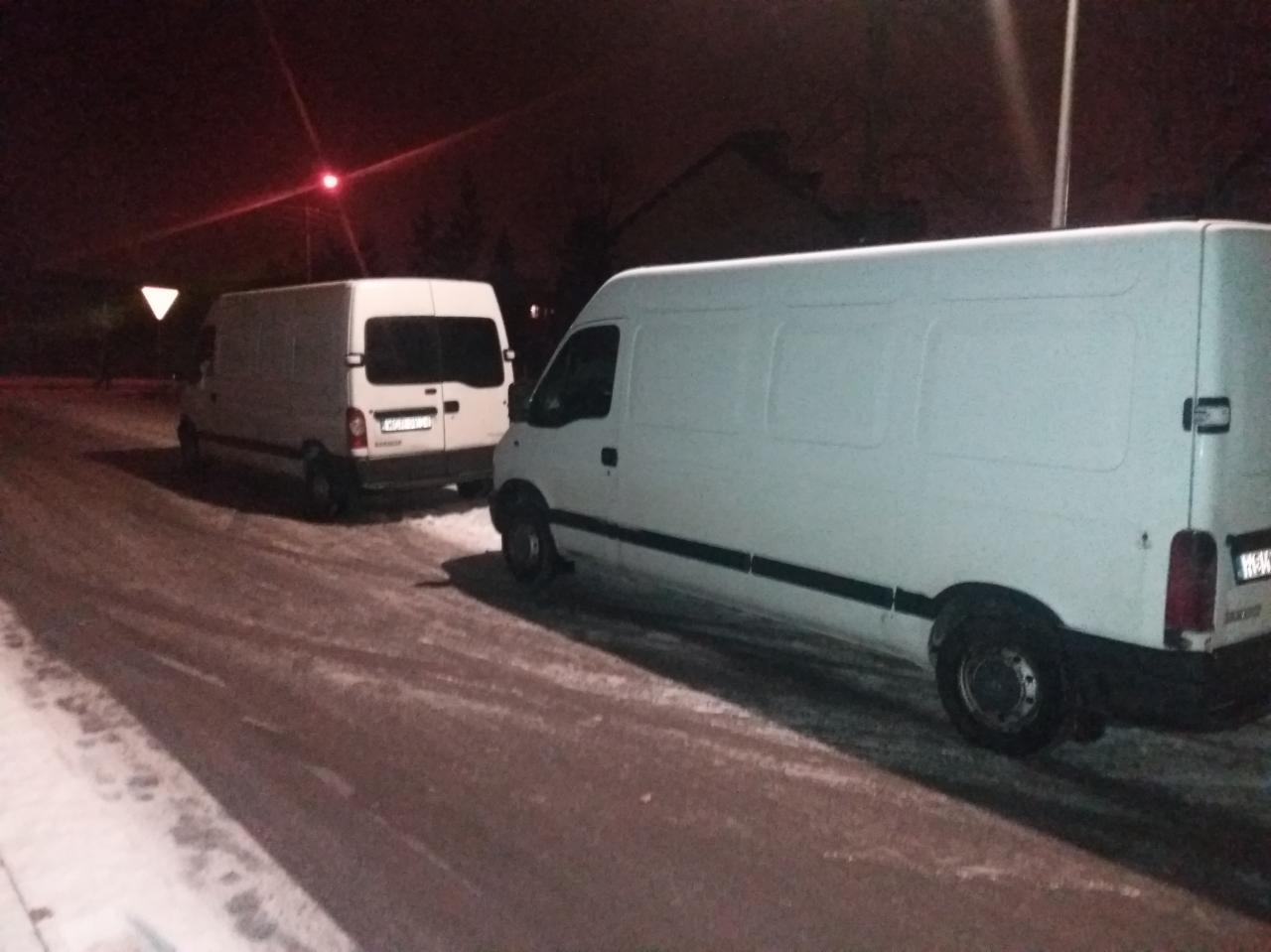 Dwa białe busy zaparkowane na ośnieżonej ulicy w Tłuszczu, nocne ujęcie z widoczną sygnalizacją świetlną w tle.