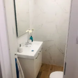 Ukończona łazienka z białą umywalką z szafką, lustrem, kwiatami w niebieskim wazonie, białymi płytkami imitującymi marmur na ścianach i drewnopodobną podłogą