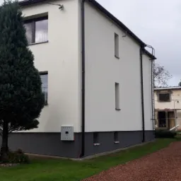 Odnowiona elewacja dwukondygnacyjnego domu z szarą podmurówką, widoczna skrzynka elektryczna na ścianie, obok rośnie wysoki iglak, w tle budynek w trakcie prac wykończeniowych i zaparkowany bus.