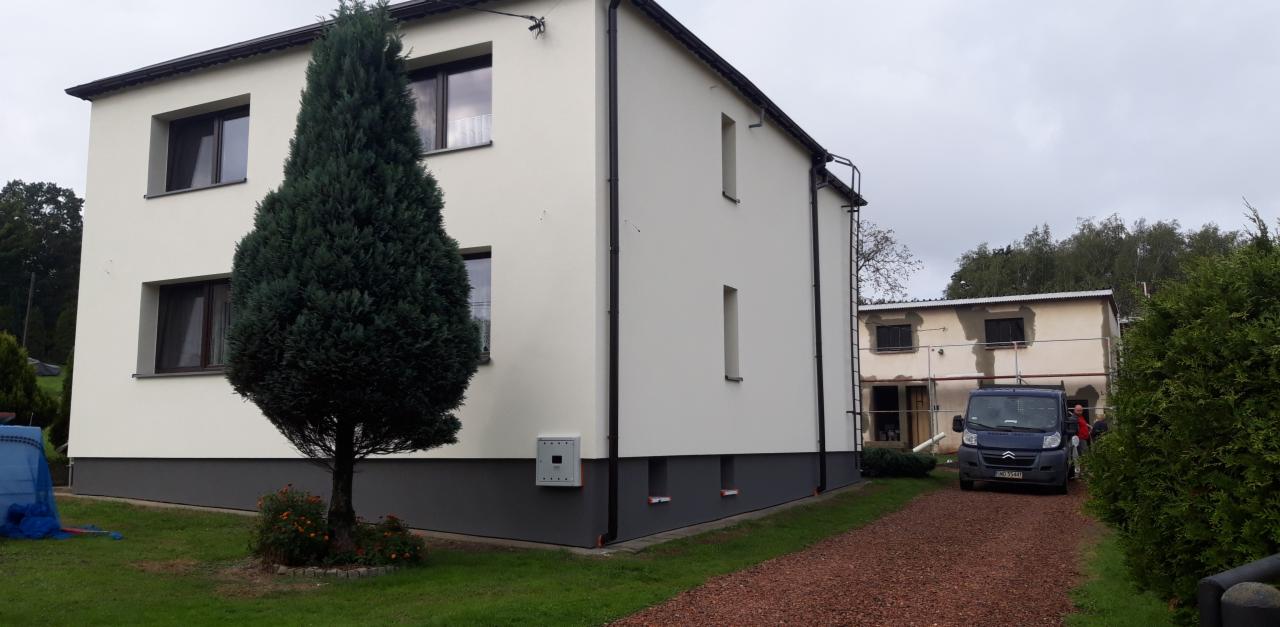 Odnowiona elewacja dwukondygnacyjnego domu z szarą podmurówką, widoczna skrzynka elektryczna na ścianie, obok rośnie wysoki iglak, w tle budynek w trakcie prac wykończeniowych i zaparkowany bus.