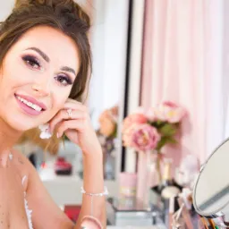 Uśmiechnięta kobieta z perfekcyjnym makijażem, w białej koronkowej sukience, dotyka ucha ozdobionego kwiatowymi kolczykami, w tle toaletka z lustrem, pędzle do makijażu i bukiet różowych kwiatów.