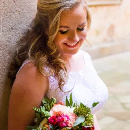 Uśmiechnięta panna młoda w koronkowej sukni, trzymająca kolorowy bukiet kwiatów, oparta o kamienną ścianę w tle okno.