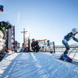 Narciarz w kasku i goglach tuż przed startem zawodów, z widocznym sprzętem pomiarowym i operatorem kamery w tle, na tle zaśnieżonych gór.