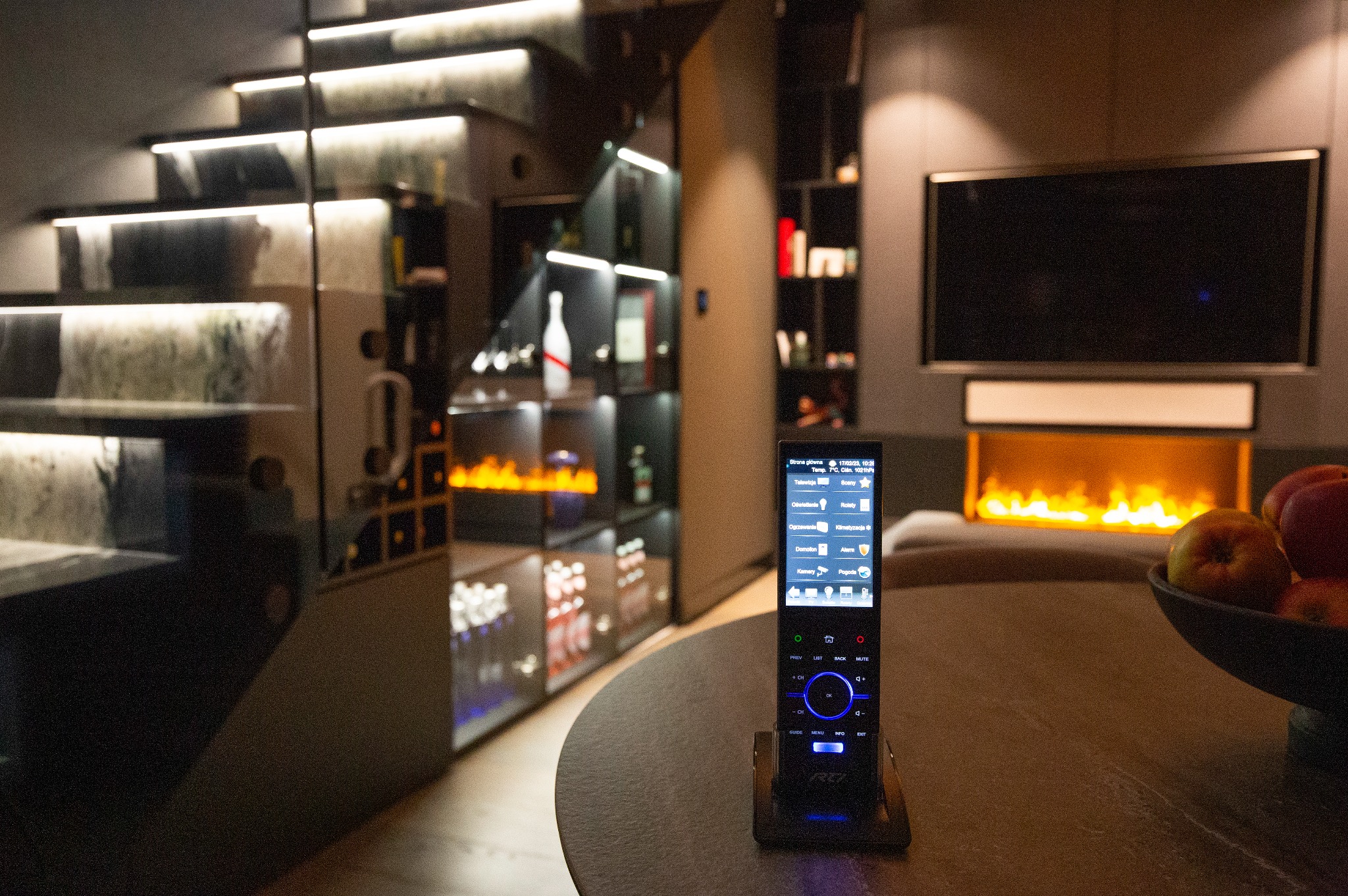 Nowoczesne wnętrze z inteligentnym systemem sterowania, na pierwszym planie pilot do zarządzania domem, w tle schody z podświetleniem LED, zabudowana szafa z podświetlonymi półkami i imitacja...