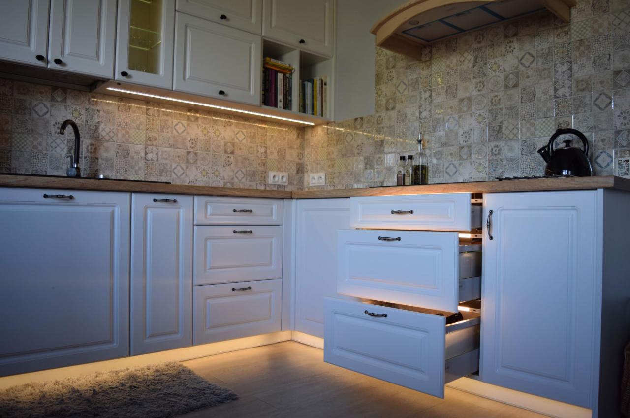 Kuchnia w stylu klasycznym z białymi szafkami, drewnianym blatem i oświetleniem LED pod szafkami i w otwartych szufladach, na ścianie dekoracyjne płytki.