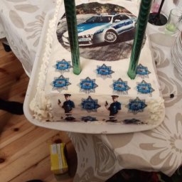 Mąż i Żona - Prostokątny tort urodzinowy z kremem, dekorowany jadalnym papierem z wizerunkiem radiowozu policyjnego i wizerunkami policjantów, ozdobiony świeczkami w kształcie cyfr '1' i '8'.