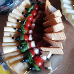 Mąż i Żona - Deska serów i przekąsek z pomidorkami cherry, bazylią, oliwkami, mini mozzarellą i krakersami na białym talerzu. Ser żółty w plasterkach na szaszłykach.