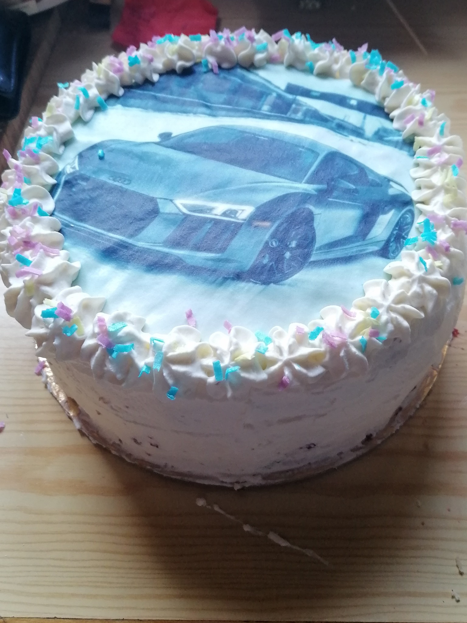 Okrągły tort z białym kremem i posypką, dekorowany jadalnym nadrukiem przedstawiającym sportowy samochód.