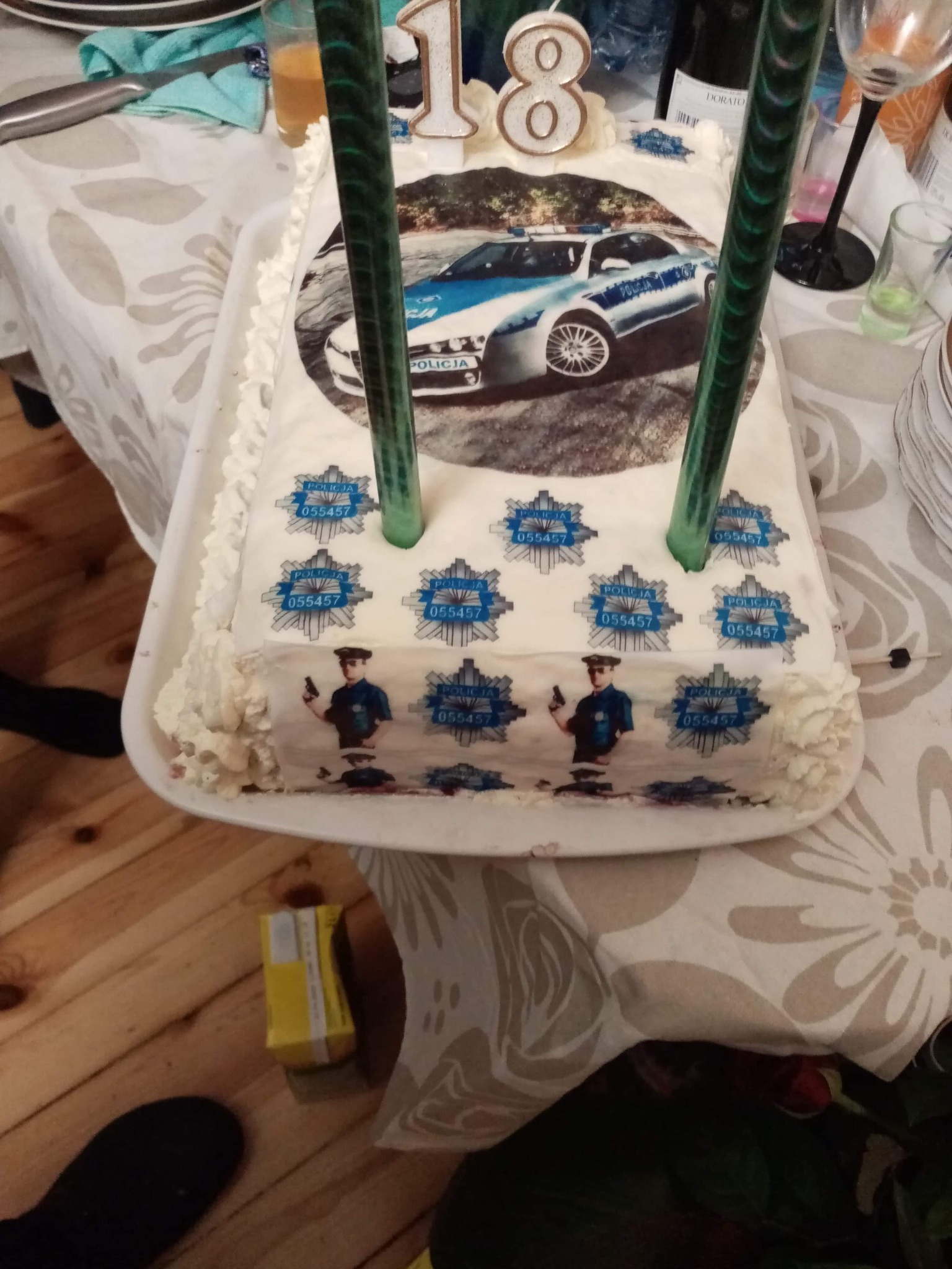 Prostokątny tort urodzinowy z kremem, dekorowany jadalnym papierem z wizerunkiem radiowozu policyjnego i wizerunkami policjantów, ozdobiony świeczkami w kształcie cyfr '1' i '8'.