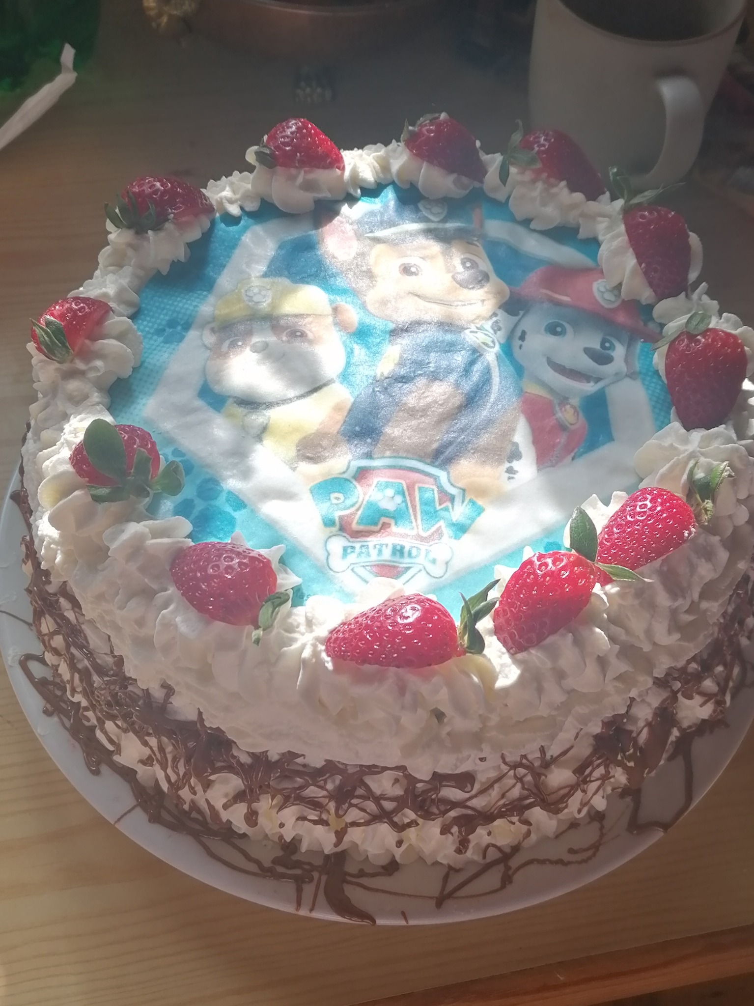 Tort urodzinowy z motywem Psiego Patrolu, udekorowany bitą śmietaną i świeżymi truskawkami, czekoladowe zdobienia na bokach, na drewnianym stole.