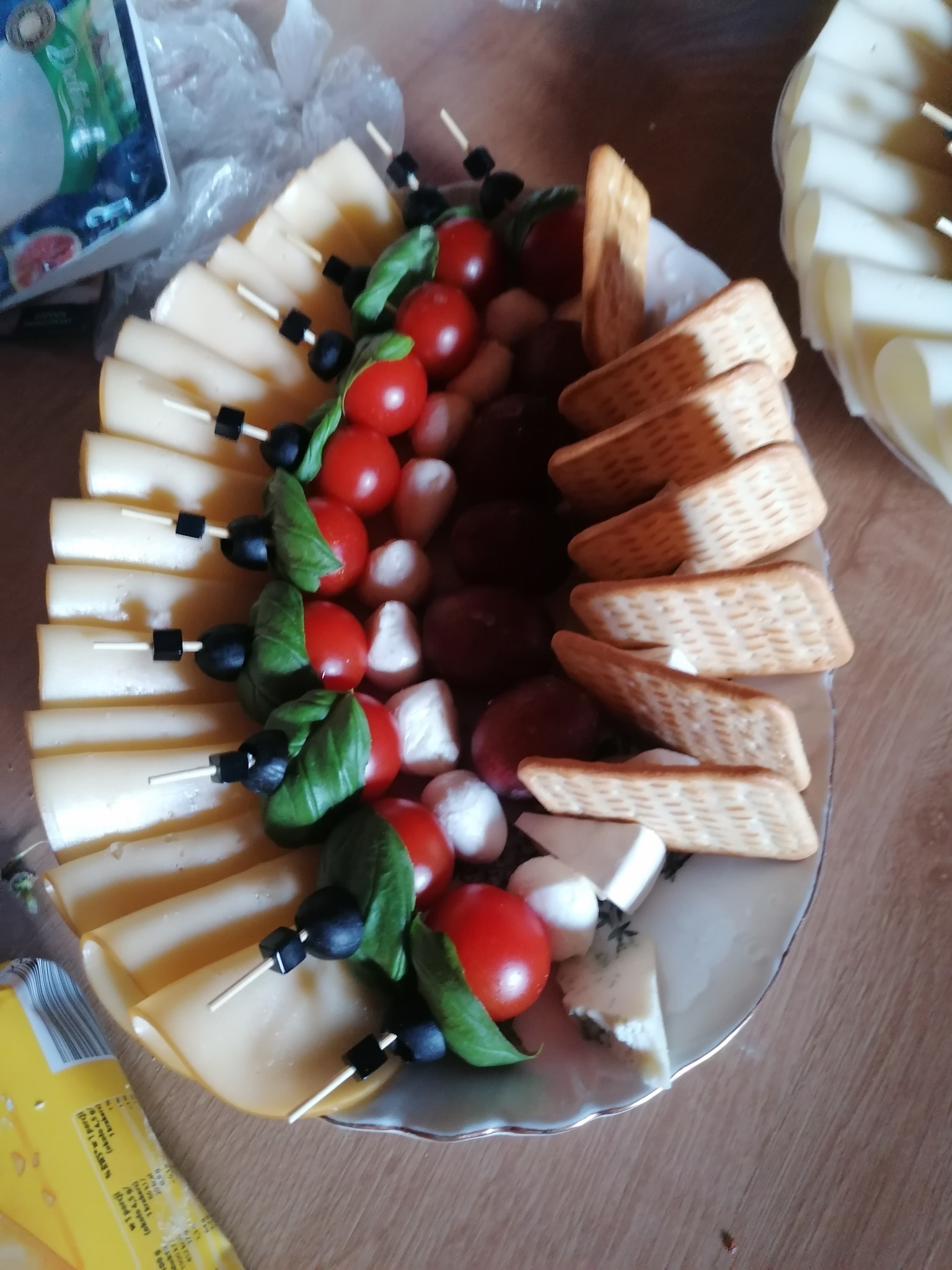 Deska serów i przekąsek z pomidorkami cherry, bazylią, oliwkami, mini mozzarellą i krakersami na białym talerzu. Ser żółty w plasterkach na szaszłykach.