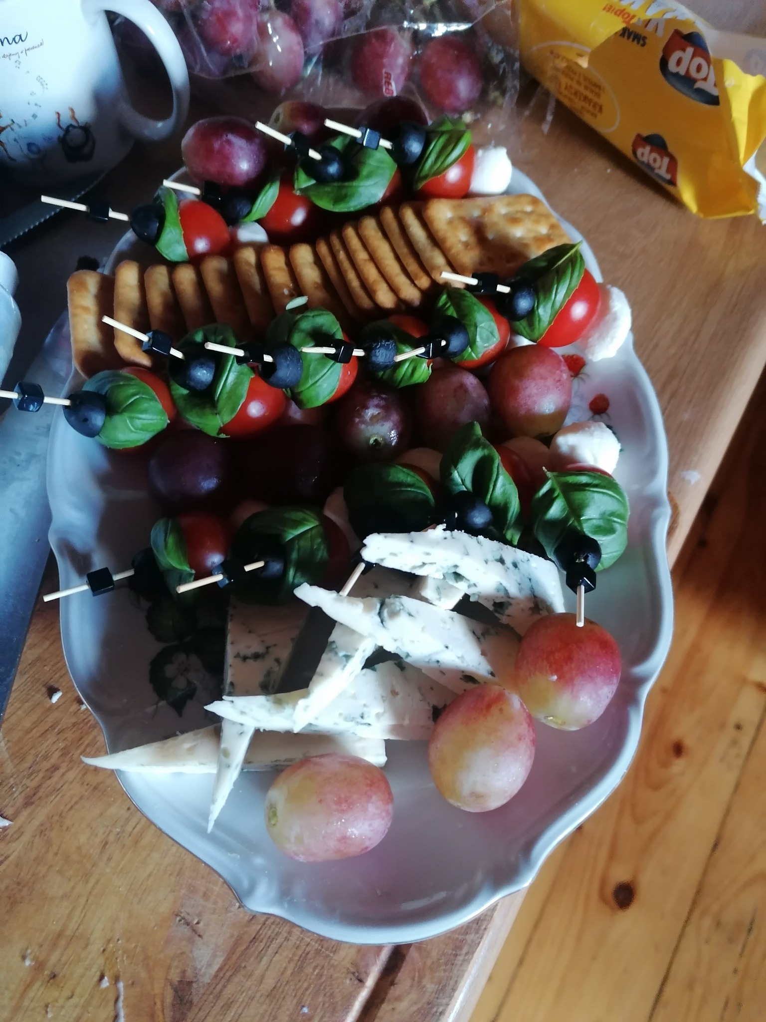 Dekoracyjna taca z przekąskami: szaszłyki z pomidorków koktajlowych, oliwek i bazylii, kostki sera pleśniowego, winogrona oraz krakersy na drewnianym stole, obok paczka krakersów.