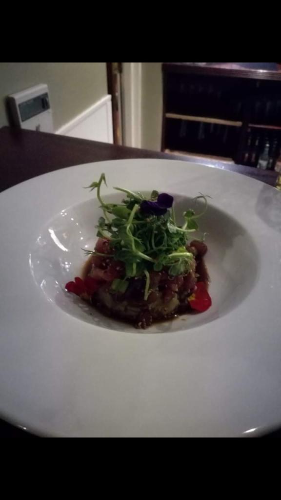 Elegancko podane danie: tatar wołowy z mikro-ziołami i jadalnymi kwiatami na białym talerzu w restauracyjnym otoczeniu.