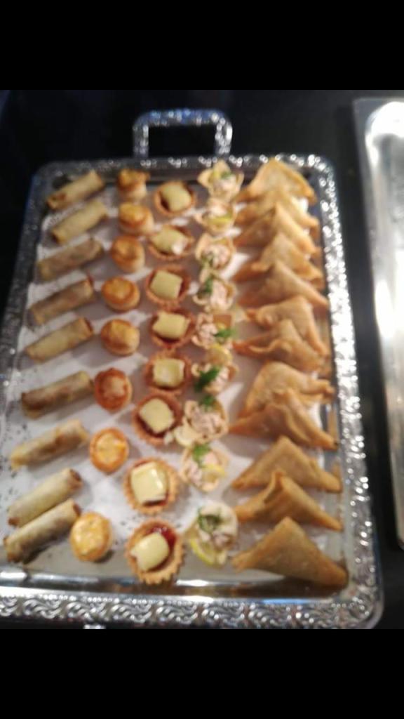 Srebrna taca z różnorodnymi przekąskami: roladki, babeczki z serem i dżemem, sałatkowe tartaletki i trójkątne pierożki.