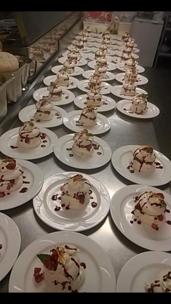 Rząd białych talerzy z pojedynczymi porcjami bezowej wieżyczki z kremem, truskawkami i polewą czekoladową, przygotowanych na metalowym blacie kuchennym.