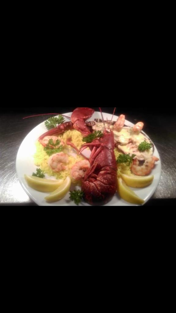 Dekoracyjnie podany talerz z homarami, krewetkami, ryżem barwionym kurkumą, cytryną i świeżą natką pietruszki na ciemnym tle.