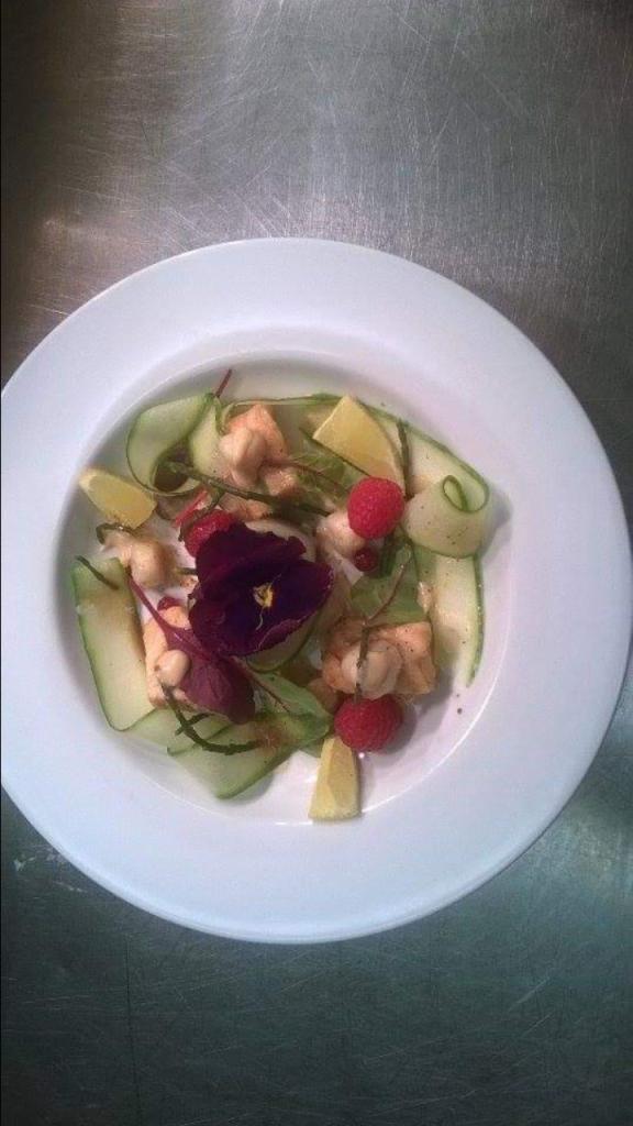 Elegancko podana sałatka z plasterkami ogórka, malinami, kawałkami cytryny i jadalnym fioletowym kwiatem na białym talerzu, na tle metalowego blatu.