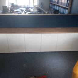 Royal Meble - Długa, niska szafka z białymi frontami i drewnianym blatem, widoczna w biurze z odbliciem regałów i biurek w tle.