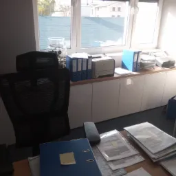 Biuro z białymi szafkami pod blatem, krzesłem z siatki, biurkiem z dokumentami i widokiem na zewnątrz przez okno z zasłoniętą budową.