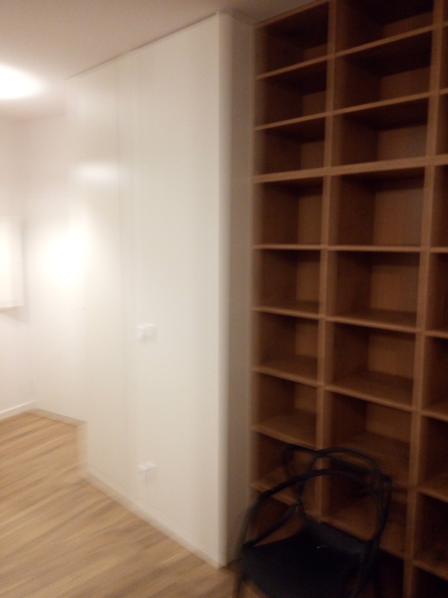 Wysoka, drewniana, otwarta półka z kwadratowymi przegródkami, przylegająca do białej ściany zabudowy w salonie, z widocznym fragmentem podłogi i krzesłem w dolnym rogu.