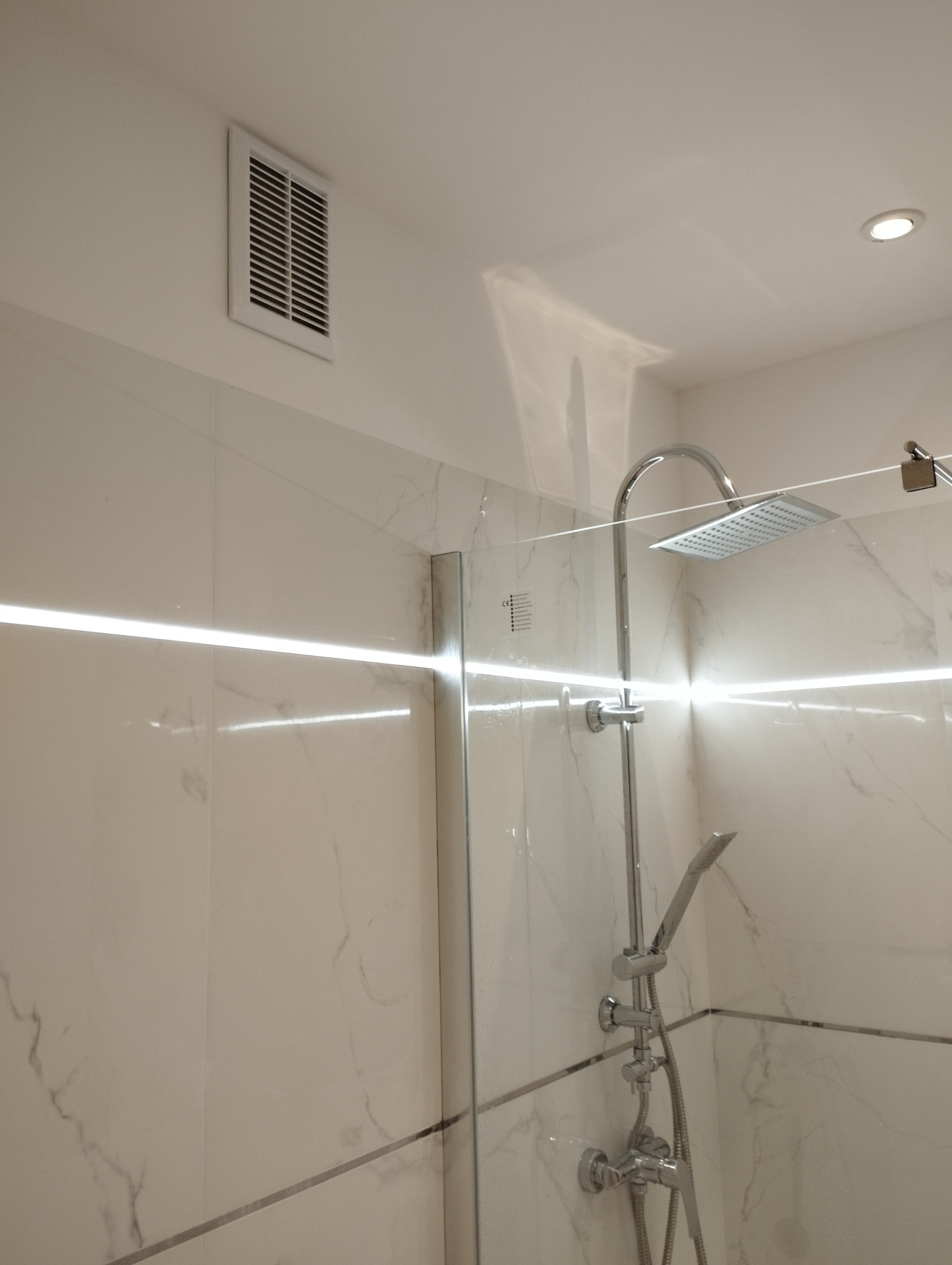 Nowoczesna kabina prysznicowa z marmurowymi płytkami, oświetleniem LED i deszczownicą. Widoczny fragment kratki wentylacyjnej w górnym rogu.