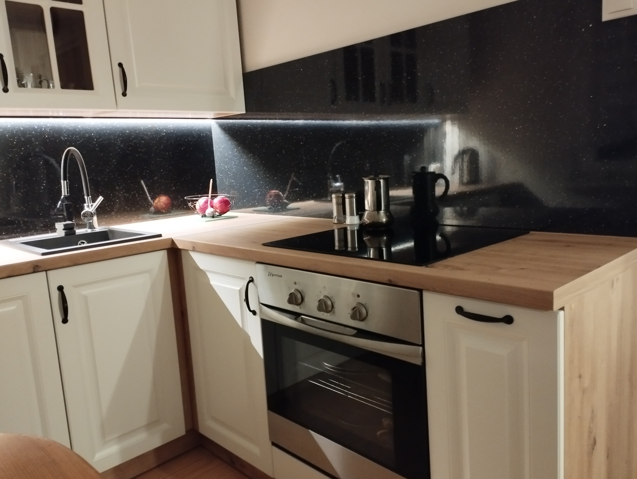 Jasna kuchnia z czarnym, błyszczącym panelem nad blatem. Widoczny zlew, płyta grzewcza i piekarnik. Blat drewniany, szafki białe z ciemnymi uchwytami.