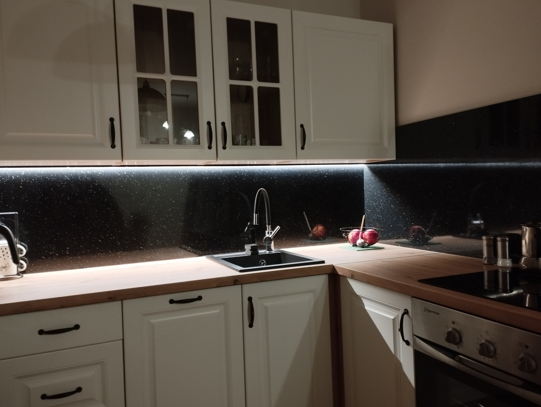 Biała zabudowa kuchenna z czarnym blatem, oświetleniem LED i połyskującym panelem ściennym. Widoczny zlew, bateria i akcesoria kuchenne. Minimalistyczny design.