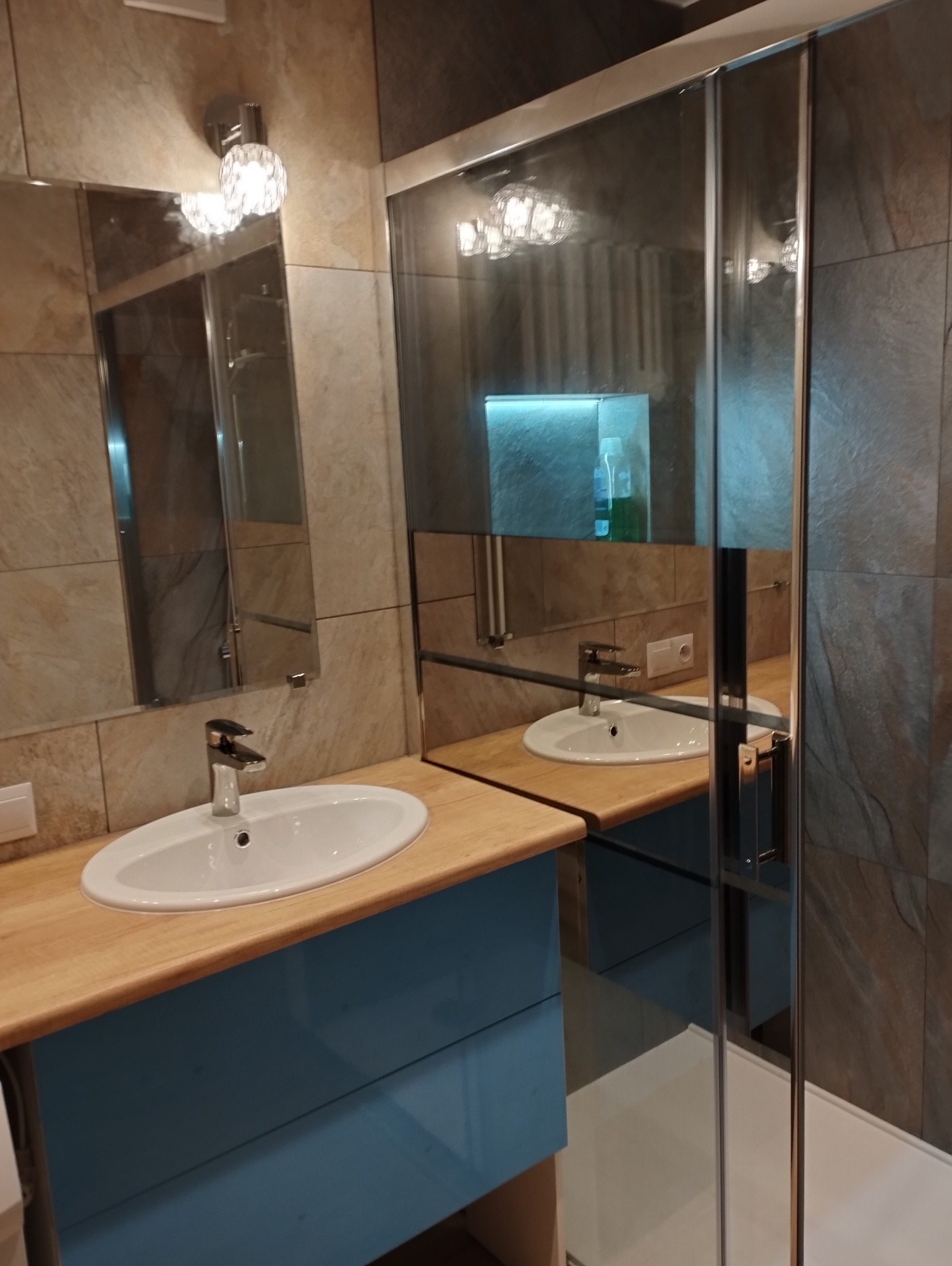 Wykończona łazienka z umywalką na drewnianym blacie i kabiną prysznicową. Ściany wyłożone płytkami imitującymi kamień. Lustro odbija oświetlenie.