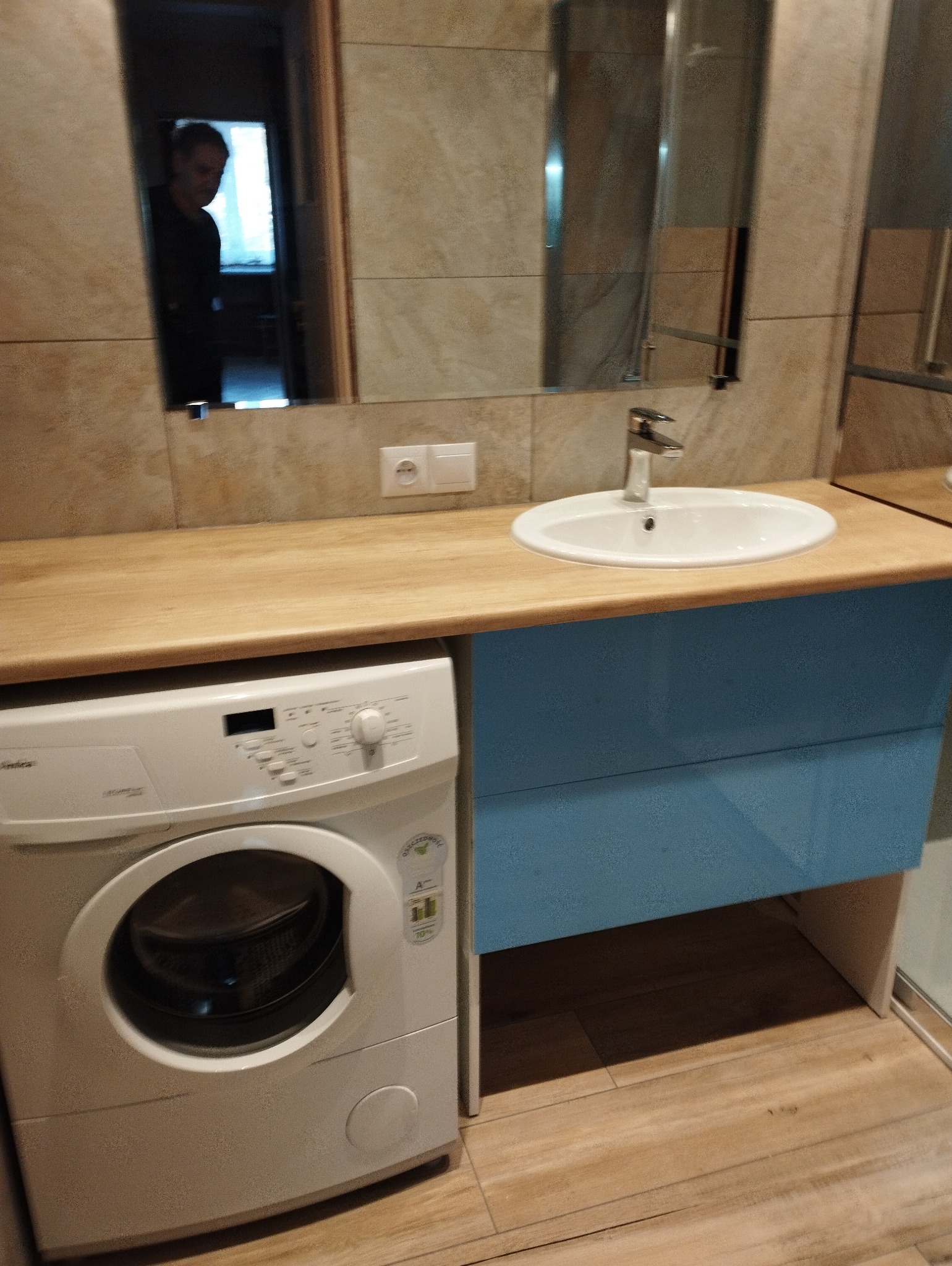 Jasna łazienka z beżowymi płytkami i drewnianym blatem. Pod blatem niebieska szafka z umywalką i pralka. W lustrze odbija się osoba.