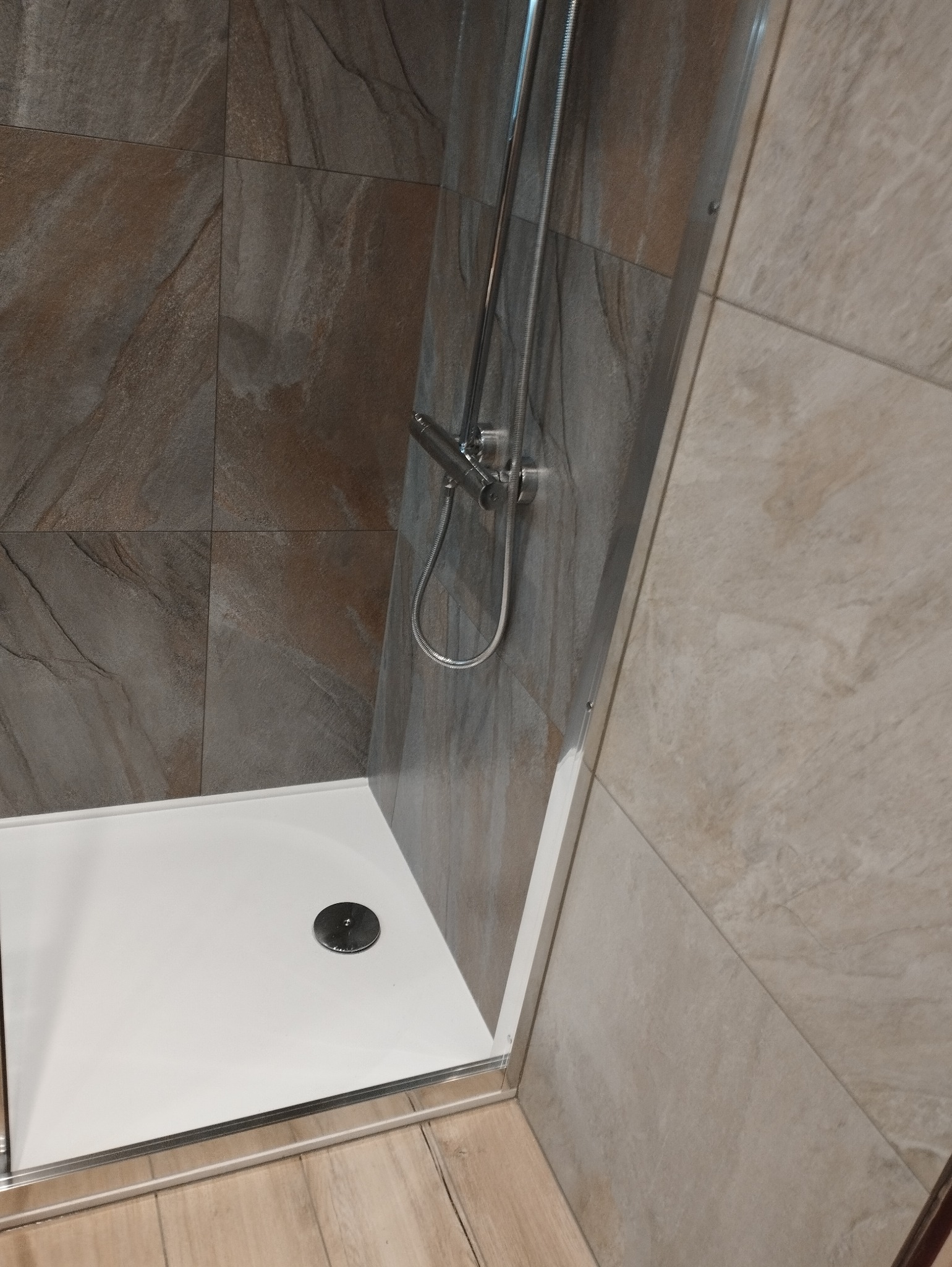 Kabina prysznicowa z odpływem liniowym i baterią, wykończona płytkami w odcieniach brązu i beżu. Widoczny brodzik i elementy armatury.