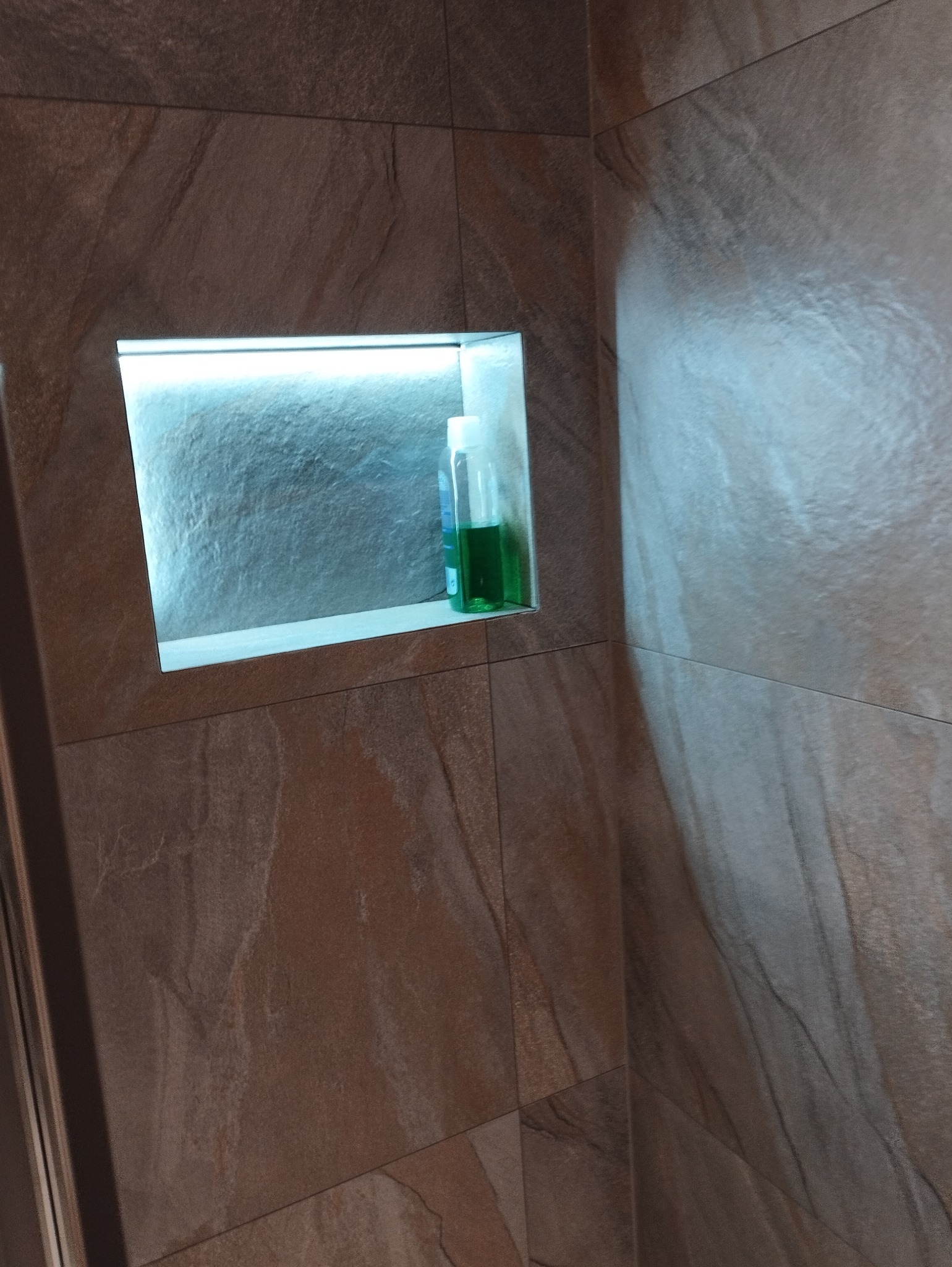 Narożnik łazienki z półką w ścianie wyłożonej brązowymi płytkami imitującymi kamień. W półce stoją dwie butelki z płynem. Półka podświetlona.